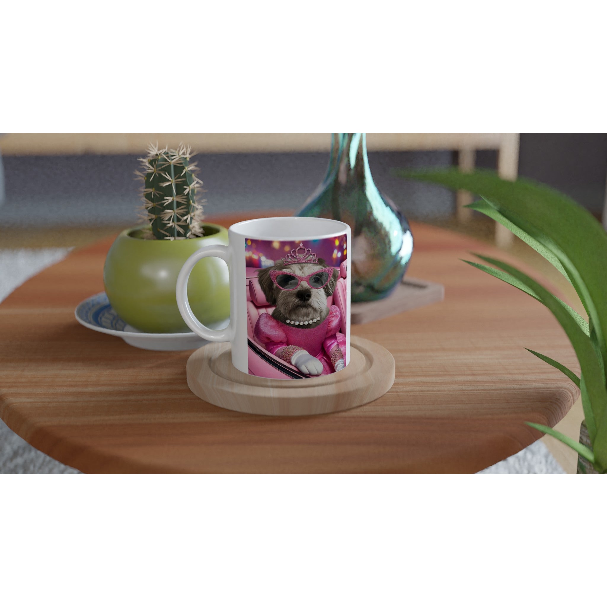 Mug blanc posé sur une table basse en bois, imprimé d’un petit chien déguisé en princesse rose avec diadème, lunettes de soleil et collier de perles, à côté d’un cactus en pot et d’un vase décoratif.