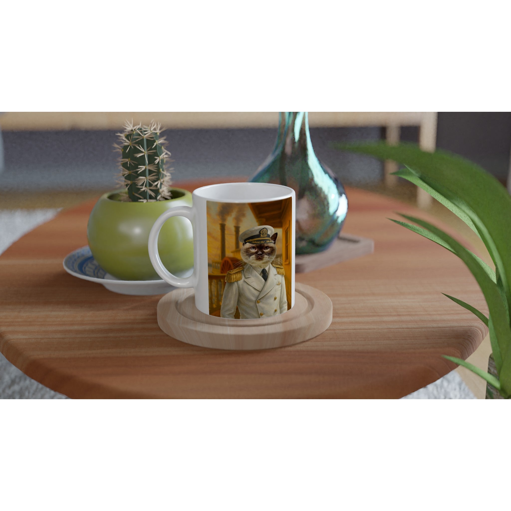 Tasse blanche posée sur un sous-verre en bois, imprimée d’un chat en uniforme de capitaine de marine, sur une table en bois avec en arrière-plan un pot vert contenant un cactus et un vase en verre coloré.