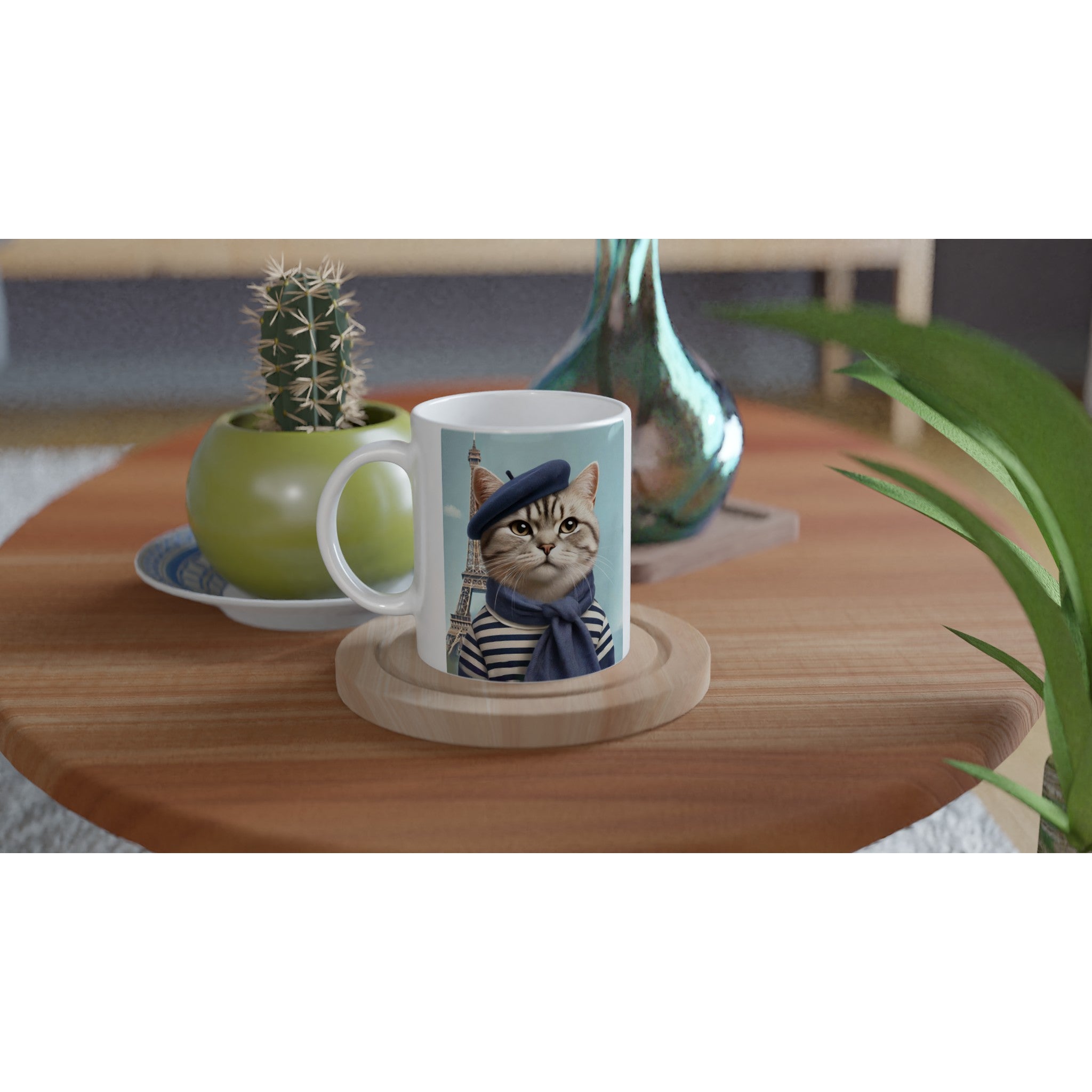 Mug en céramique blanche orné d’un chat portant un béret bleu et une marinière devant la tour Eiffel, posé sur un sous-verre en bois au milieu d’une table en bois avec un cactus en pot et un vase décoratif.