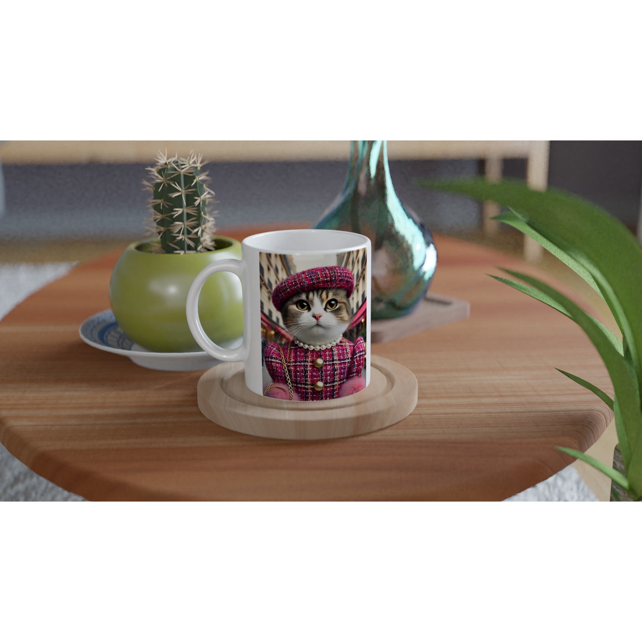 Tasse blanche posée sur une table basse en bois, illustrée d’un chat élégant vêtu d’un tailleur et d’un béret roses en tweed avec collier de perles, accompagnée en arrière-plan d’un petit cactus en pot vert et d’un vase en verre turquoise.
