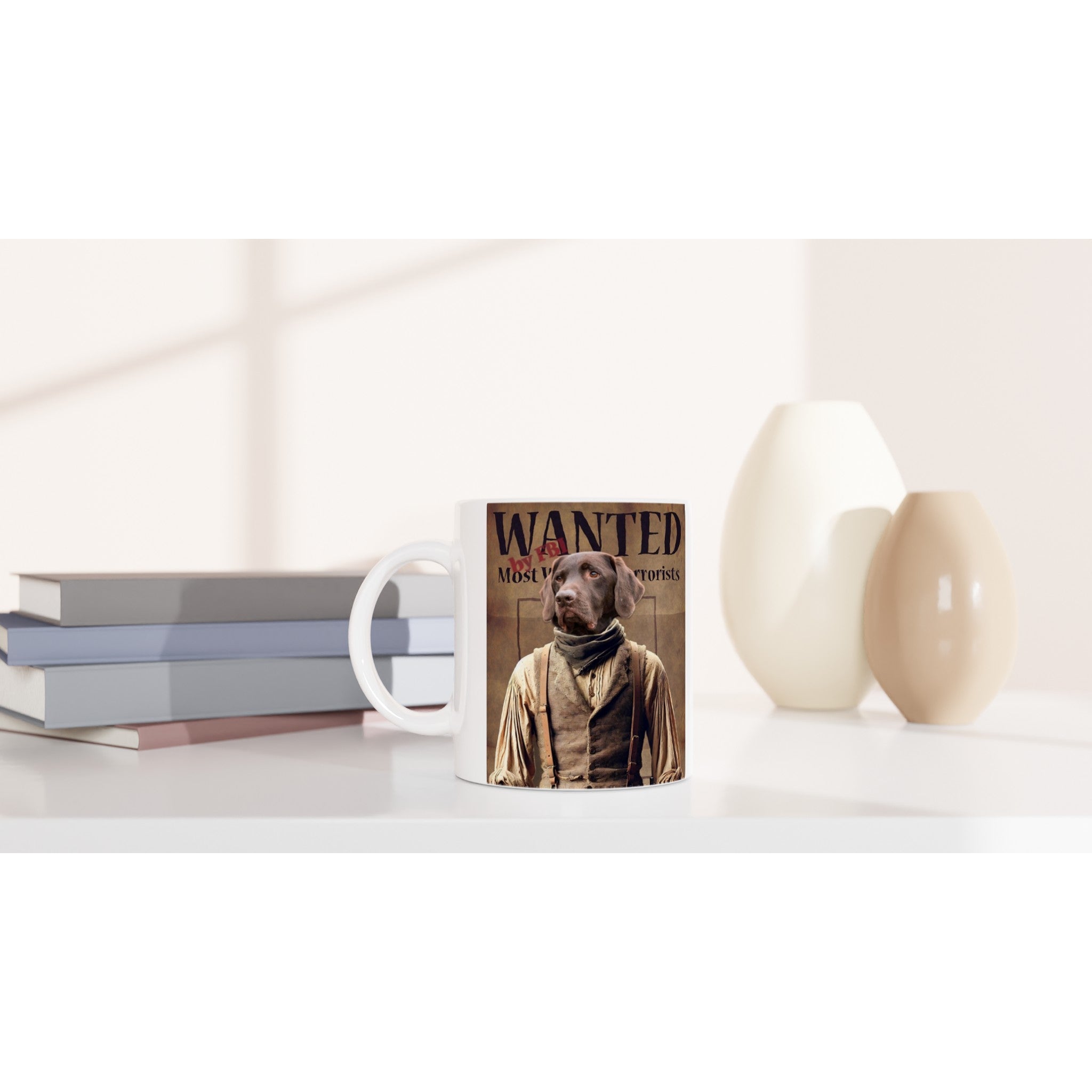 Mug blanc imprimé d’une fausse affiche “WANTED Most Wanted Terrorists” montrant un chien labrador en costume d’époque, posé sur une table à côté de livres et de vases beiges.