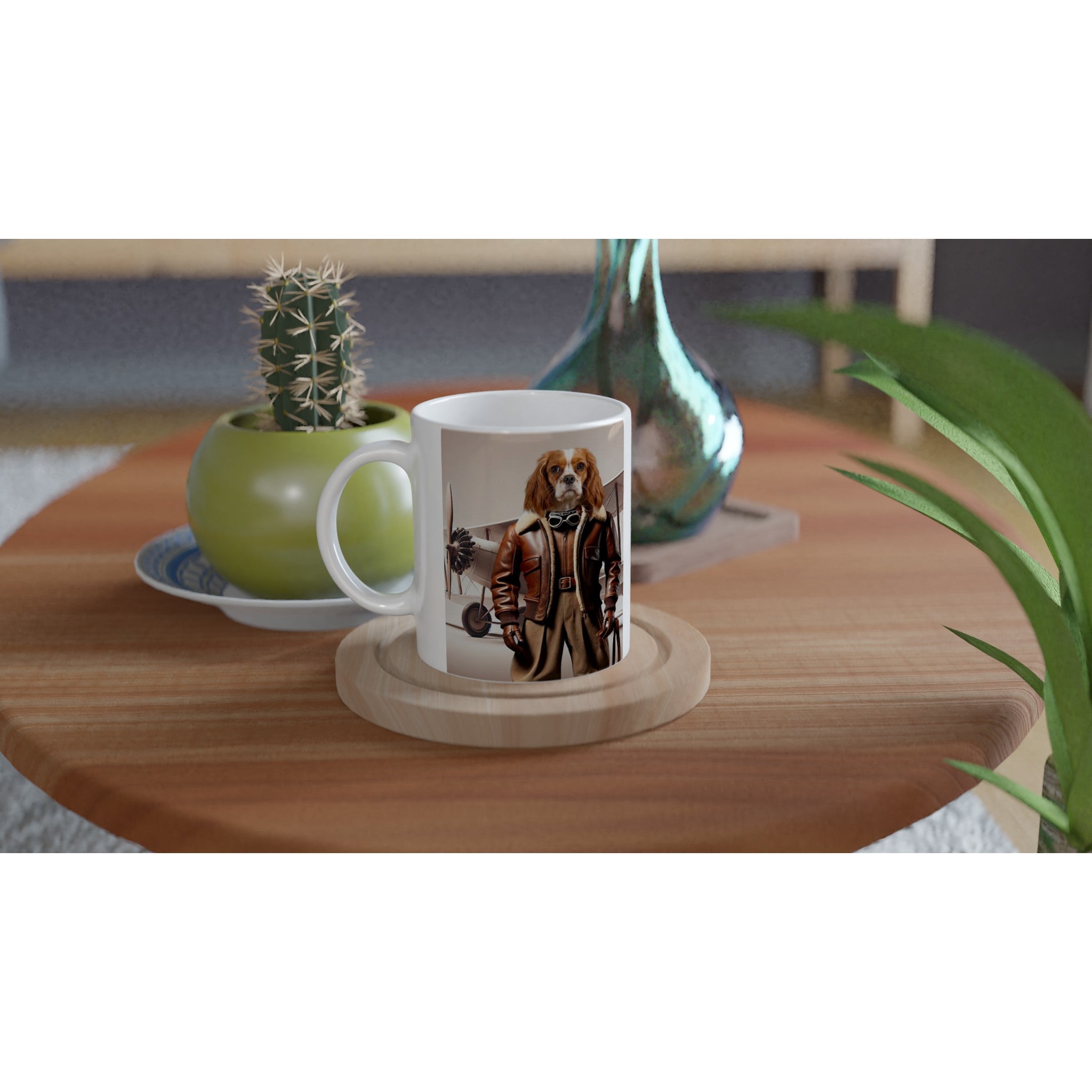 Mug blanc illustré d’un chien Cavalier King Charles en tenue d’aviateur devant un biplan, posé sur une table basse en bois à côté d’un pot de cactus vert et d’un vase décoratif