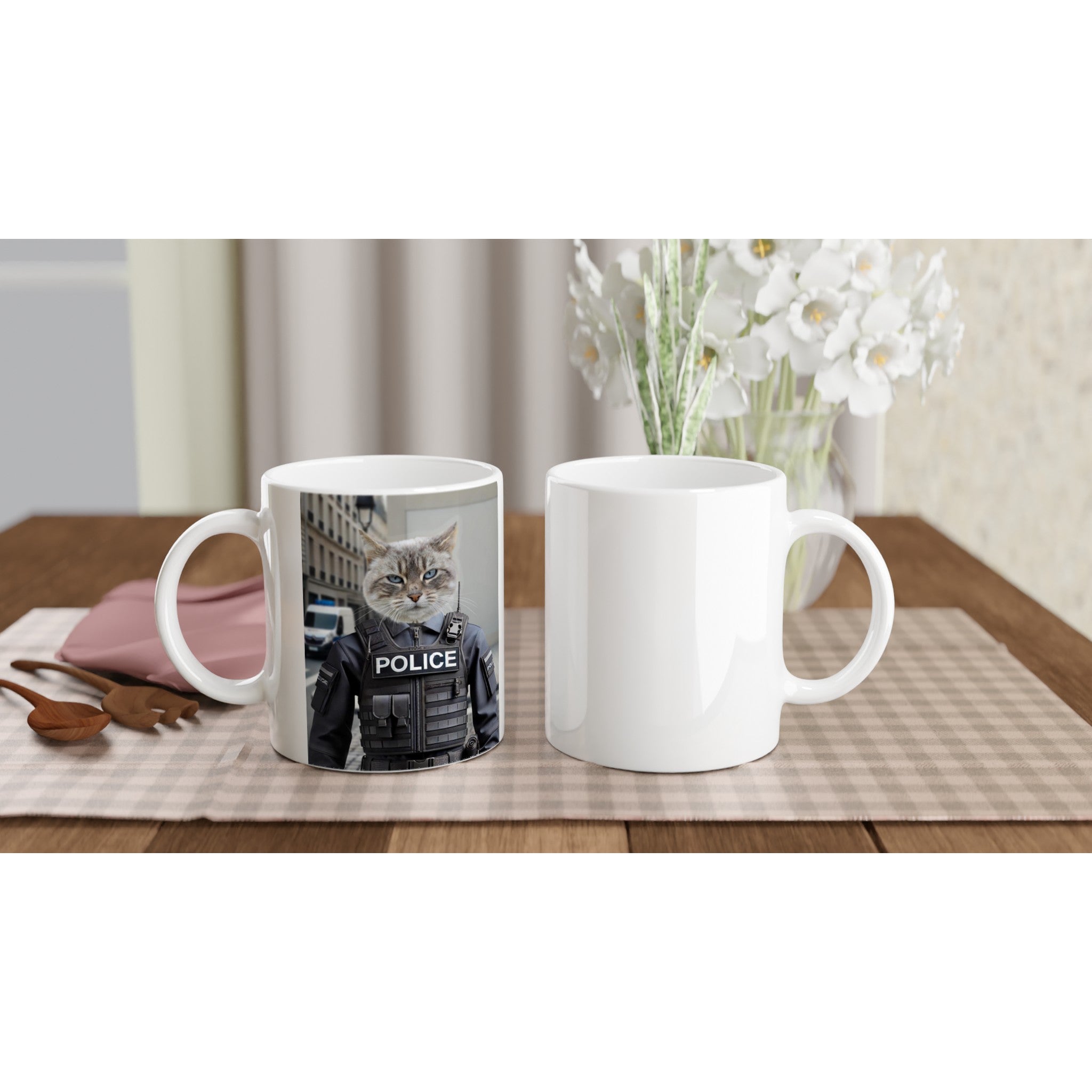 Deux mugs blancs posés sur une table en bois recouverte d’une nappe à carreaux beiges : l’un porte l’impression d’un chat en uniforme de police, l’autre est vierge, accompagnés de cuillères en bois, d’une serviette rose et d’un vase de fleurs blanches.