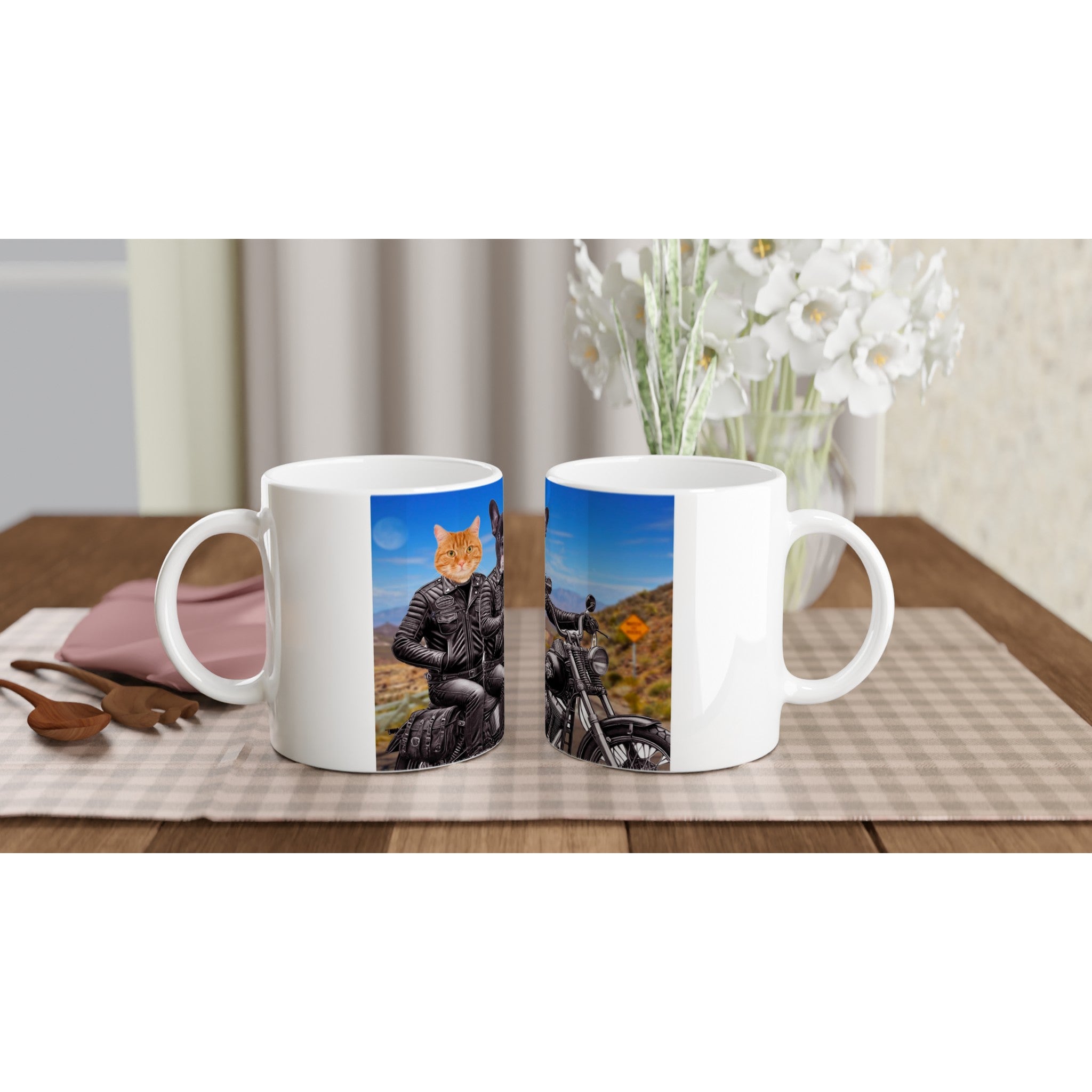 Deux mugs blancs posés sur une table à carreaux : l’un imprimé d’un chat roux en blouson de cuir pilotant une moto dans un paysage désertique, accompagné de cuillères en bois, d’une serviette rose et d’un vase de fleurs blanches.